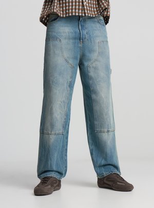 Pantaloni in denim azzurro chiaro con vestibilità ampia, caratterizzati da grandi tasche, leggera scoloritura e cuciture rinforzate. Abbinati a sneakers scure.
