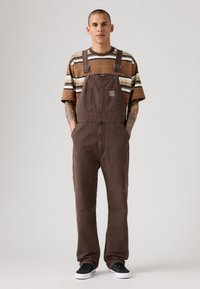 Jonge man staat met zijn handen in zijn zakken, draagt een bruine overall, een gestreept bruin en beige t-shirt, en zwarte sneakers tegen een lichte achtergrond.