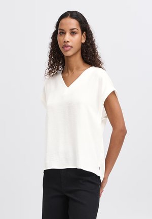 Giovane donna con capelli ricci che indossa una maglietta bianca a maniche corte con scollo a V e pantaloni neri, in piedi davanti a uno sfondo chiaro e uniforme.