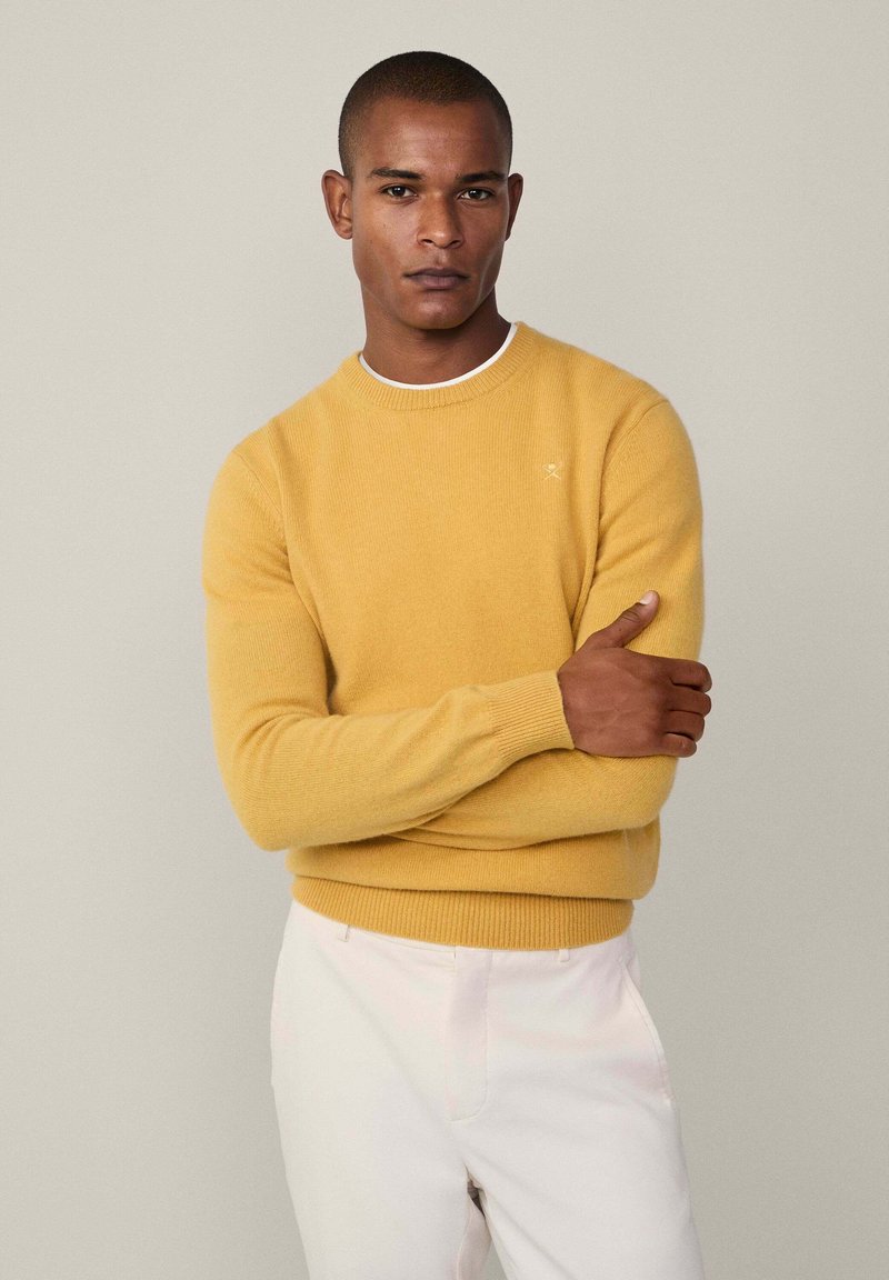 Žlutý pletený svetr s kulatým výstřihem, dlouhými rukávy a žebrovanými manžetami, v kombinaci se světle zbarvenými kalhotami. Jemný detail loga na hrudi.