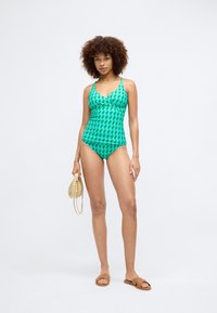 Green bikini set featuring a geometric pattern, adjustable straps, flared top, and matching bottom. Model holds a woven bag, wearing tan sandals.