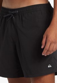 Bañador negro con cinturilla elástica, cordón ajustable, bolsillos laterales y parche de logo en la pierna izquierda. Tela lisa, sin textura.