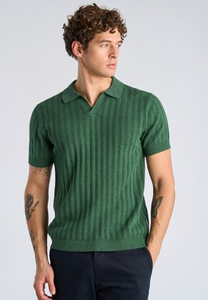 Junger Mann mit lockigem Haar, der ein grünes, kurzärmeliges, geripptes Poloshirt und dunkle Hosen trägt, blickt nach rechts und hat Tätowierungen auf beiden Armen.