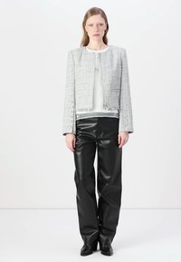 Giacca cropped in tweed grigia con dettagli in frange, bottoni metallici, indossata sopra una maglietta bianca e pantaloni in ecopelle nera. Scarpe nere completano l'outfit.