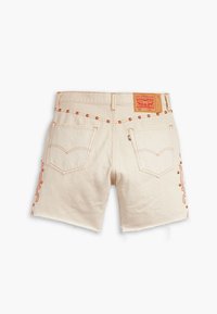 Shorts de denim beige con bajo deshilachado, costuras naranjas, bordado decorativo y acentos de clavos metálicos. La etiqueta en la parte trasera es marrón con detalles rojos.