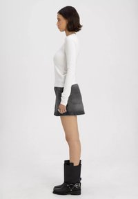 Langärmliges weißes Oberteil, schwarzer Jeansrock und kniehohe schwarze Stiefel mit Schnallenakzenten, im Profil vor einem einfarbigen Hintergrund gezeigt.