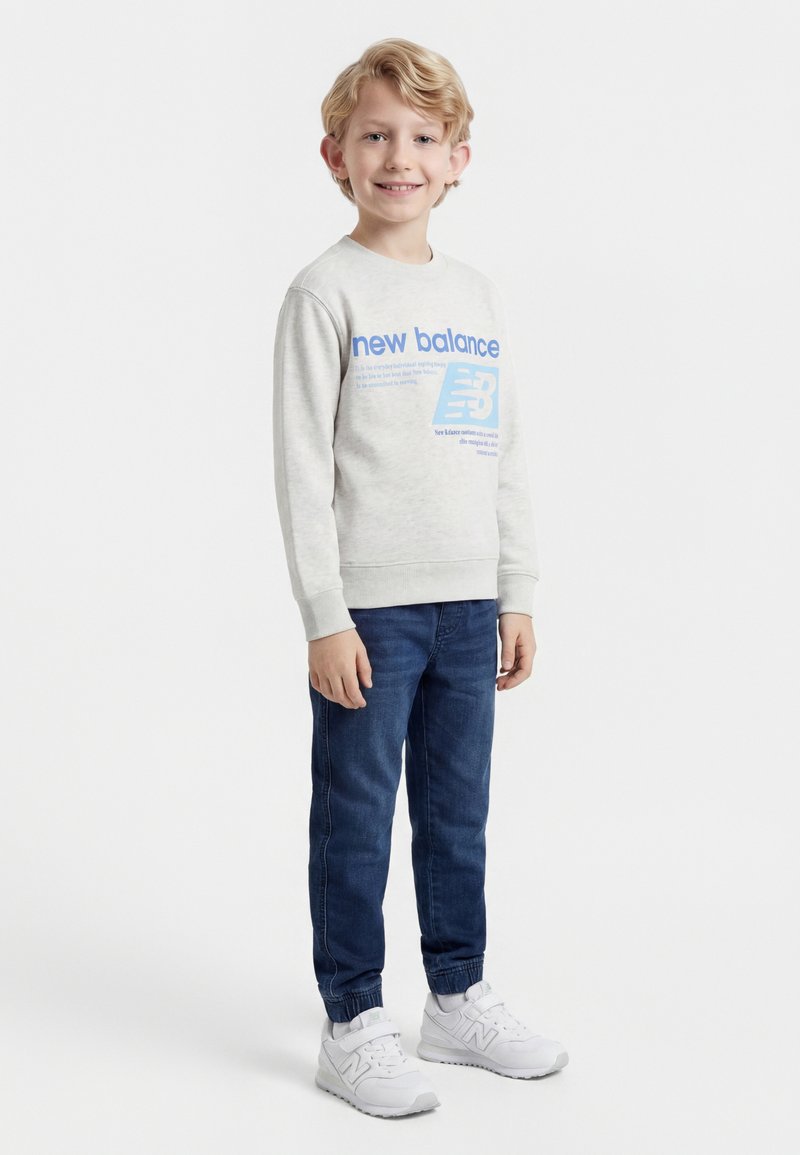 Χαμογελαστός νεαρός αγόρι που στέκεται, φοράει ανοιχτό γκρι φούτερ New Balance, μπλε τζιν και λευκά αθλητικά New Balance σε λευκό φόντο.