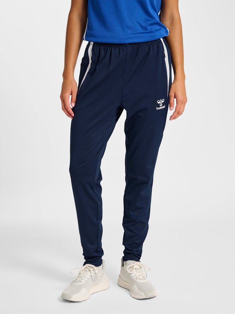 Personne portant un pantalon de sport bleu marine avec des bandes blanches sur les côtés, des baskets blanches et un t-shirt bleu, debout devant un fond uni.