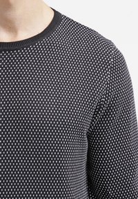 Pull noir avec un motif de tricot texturé, orné de petits triangles blancs. Modèle col rond avec un col et des poignets côtelés.