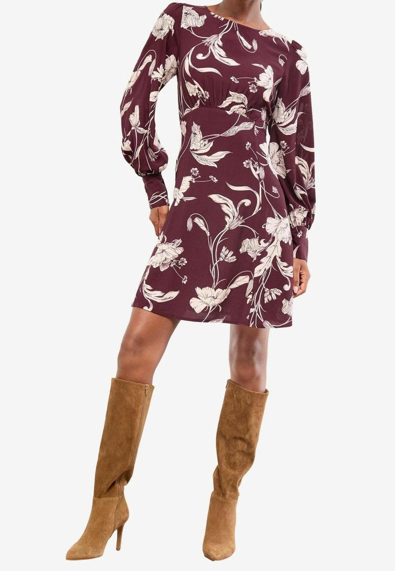 Femme portant une robe bordeaux à manches longues avec des motifs floraux blancs et des bottes hautes en daim beige à talons.