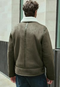 Chaqueta de ante verde oliva con un cuello de piel de oveja blanco, que presenta una costura vertical en la parte trasera y acentos de botones en los lados.