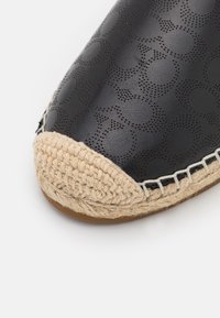 Bout en cuir noir style espadrille avec motif perforé et couture de semelle en jute tressé beige sur fond blanc.