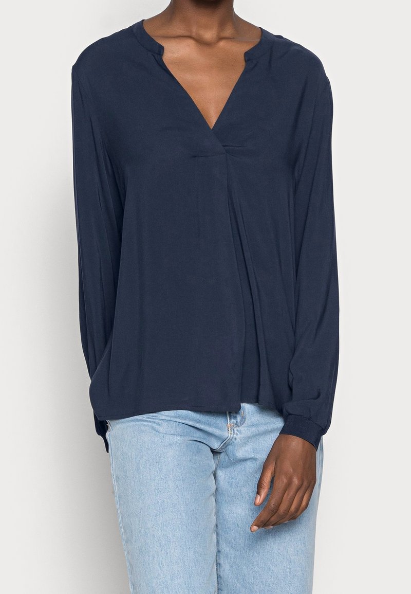Blusa navy a maniche lunghe con scollo a V, caratterizzata da una texture morbida e una parte anteriore plissettata. Abbinata a jeans azzurri chiari. Design semplice, senza motivi.