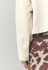 Cremefarvet denimjakke med cropped design og lomme på ærmet, parret med en brun-og-cremefarvet mønstret nederdel. Struktureret stof.