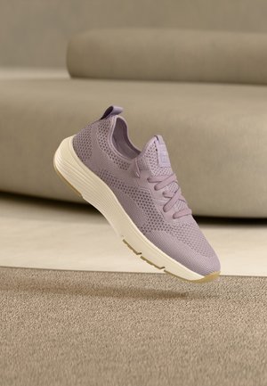 Baskets en maille violet clair avec semelle blanche épaisse flottant au-dessus d'un tapis beige, canapé beige en arrière-plan.