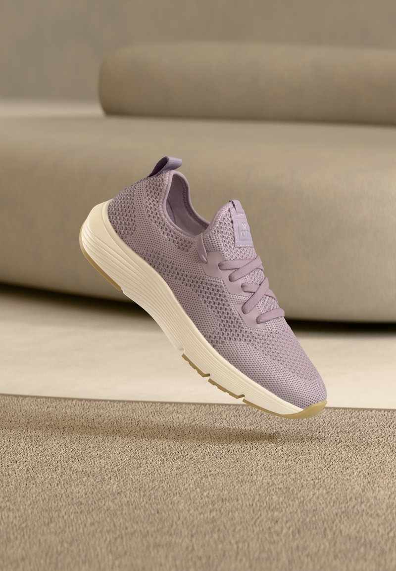 Ανοιχτό μωβ πλεκτό αθλητικό παπούτσι με παχύ λευκό πάτο που αιωρείται πάνω από μπεζ χαλί, με μπεζ καναπέ στο παρασκήνιο.