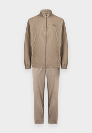 Beige Reißverschlussjacke mit elastischen Bündchen und passende Hose, beide mit kleinen EA7 Emporio Armani Logos auf Brust und Oberschenkel.