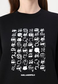 Svart hettegenser med en hvit grafikk av 64 stiliserte ansiktsillustrasjoner, inkludert solbriller, bart og ulike ansiktsuttrykk.