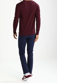 Bordeauxrotes Langarmhemd kombiniert mit navyblauen Hosen und weißen Turnschuhen. Das Hemd hat ein enganliegendes Design und eine weiche Textur, mit einem runden Ausschnitt.