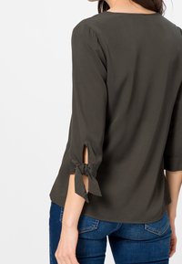 Dunkelgrüne Bluse mit drei Viertelärmeln, die mit Bindebändern an den Bündchen, einem runden Halsausschnitt und einer glatten Textur ausgestattet ist. Getragen mit blauen Jeans.