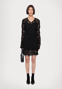 Abito in pizzo nero con maniche lunghe, caratterizzato da motivi intricati e dettagli di perline. Taglio a metà coscia con orlo scallop. La modella tiene una borsa nera.