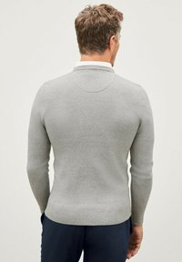 Pull en maille gris avec un design côtelé, coupe ajustée et petit col, porté sur un pantalon sombre. Détails de couture visibles sur les épaules.