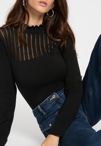 Haut noir avec un panneau transparent texturé, doté d'un col à volants ; associé à un jean en denim bleu foncé taille haute avec des accents en métal.