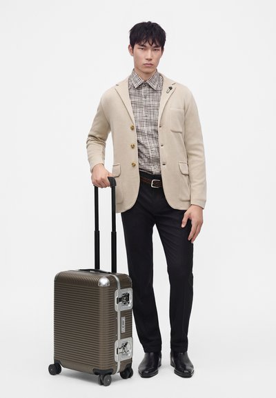 Homme portant un blazer beige et une chemise à carreaux, debout tenant la poignée d'une valise rigide marron à roulettes sur fond blanc.
