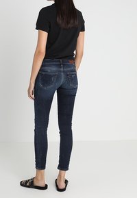 Jeans ajustados de denim azul oscuro con una textura descolorida, que cuentan con bolsillos traseros y un parche de cuero marrón en la banda de la cintura.