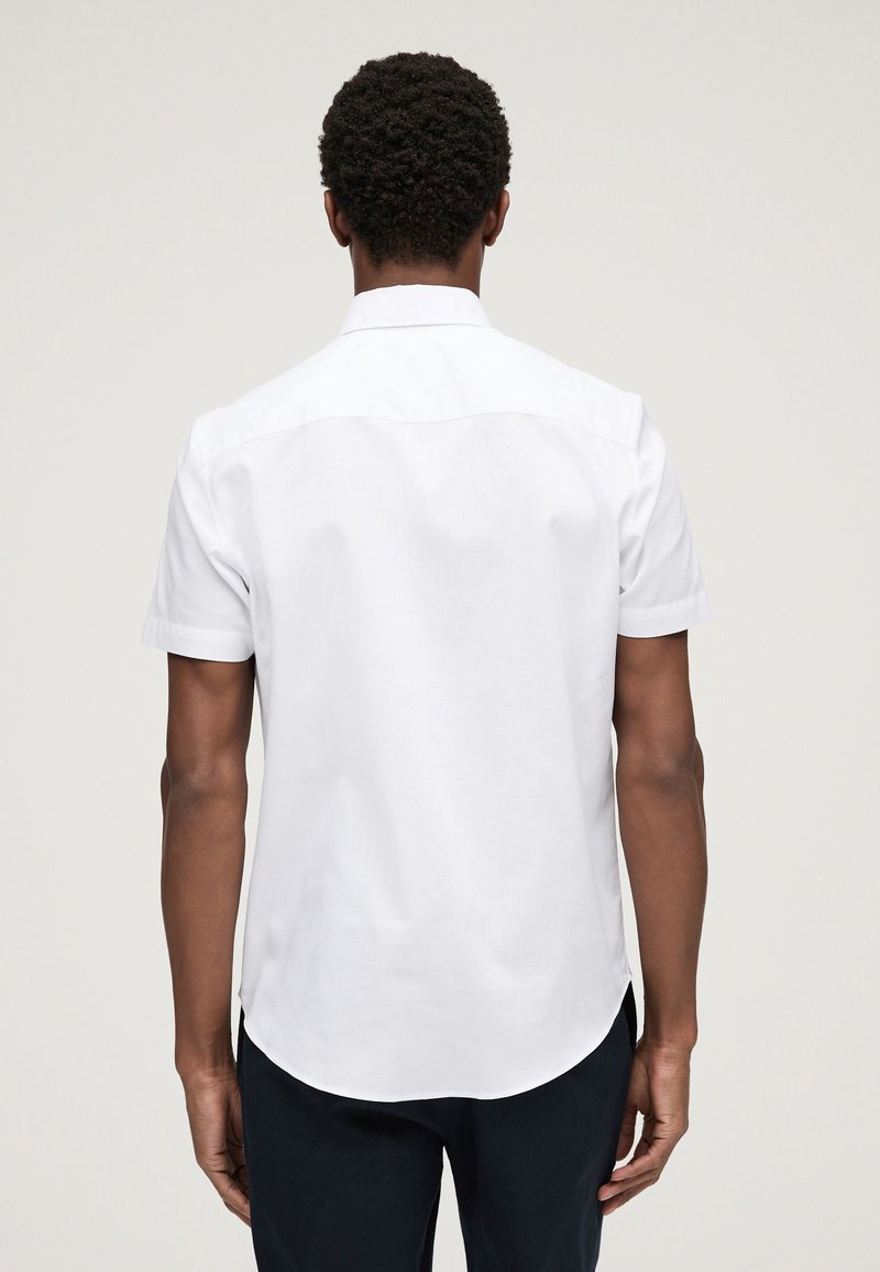 Homme aux cheveux courts et bouclés portant une chemise blanche unie à manches courtes et un pantalon foncé, debout de dos face au spectateur.