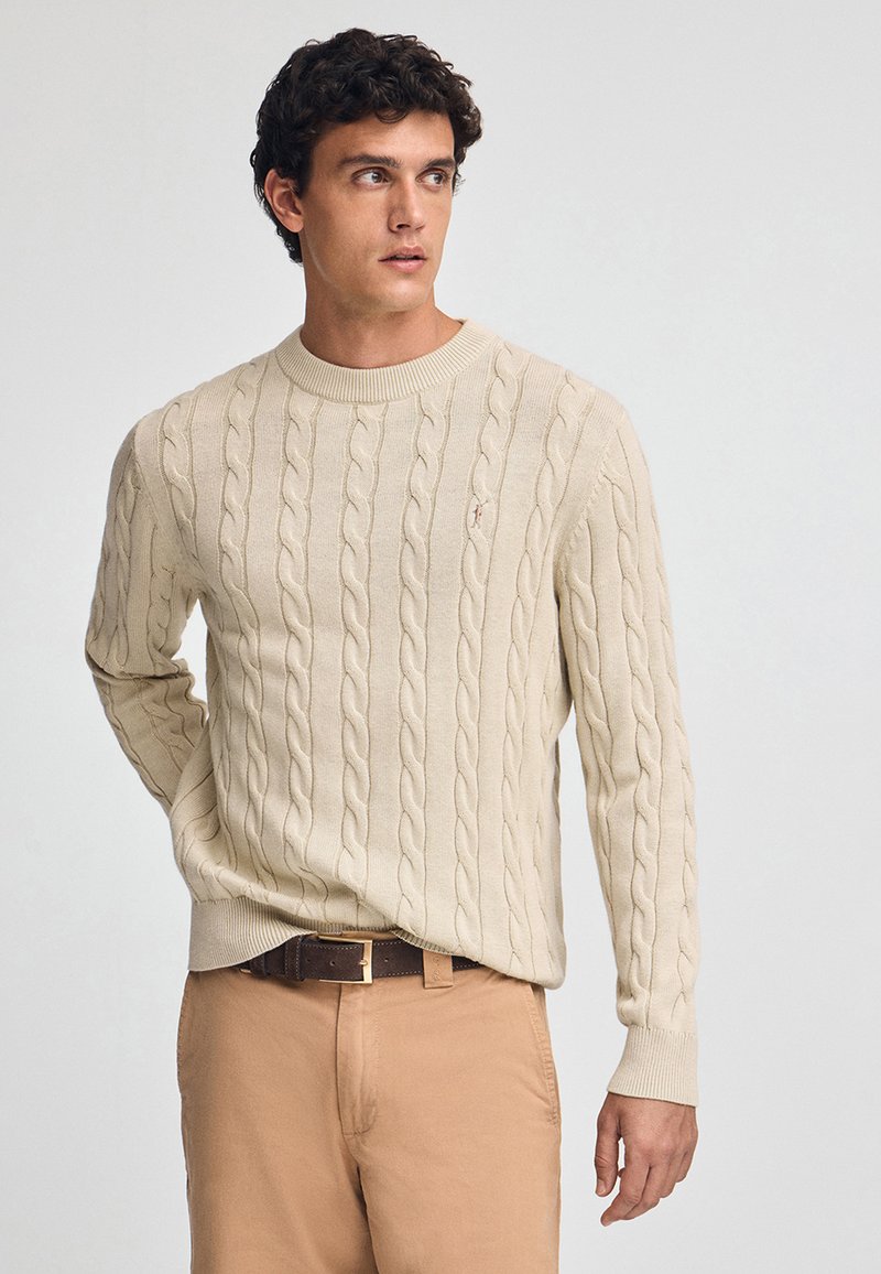 Pull en tricot beige avec un col et des poignets côtelés, arborant un petit logo sur la poitrine, associé à un pantalon marron clair et une ceinture.