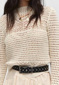 Suéter de punto beige claro con patrón calado, que presenta un cuello alto y mangas largas. Combinado con un cinturón negro con ojales de metal.