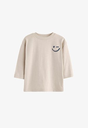 Beige katoenen t-shirt met driekwart mouwen, ronde hals en een donkerblauwe smiley-gezichtsafbeelding op de linkerkant van de borst.