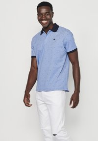 Ljusblå pikétröja med marinblå krage och manschetter, prydd med ett upprepat diamantmönster. Bärs med vita slitna jeans.