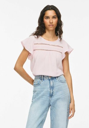 Femme aux cheveux bouclés et foncés portant un chemisier rose clair avec détail en dentelle et un jean taille haute bleu clair sur fond blanc.