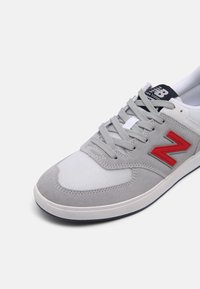 Šedé a bílé tenisky New Balance s červeným logem "N", šedými tkaničkami a tmavě modrým jazykem, zobrazené z přední strany na bílém pozadí.