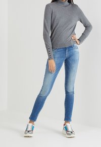 Grauer Rollkragenpullover mit langen Ärmeln und Knopfdetails, kombiniert mit blauen Skinny-Jeans und mehrfarbigen Sneakers.
