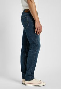 Vaqueros de denim azul con un ajuste slim, que cuentan con un parche de cuero en la parte trasera y costuras en contraste. Combinados con zapatillas de lona blancas.