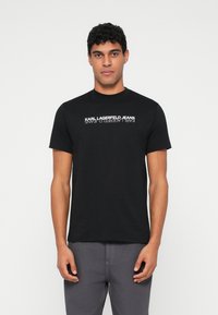 Melnas kokvilnas T-krekls ar uzrakstu "KARL LAGERFELD JEANS", kas drukāts baltā krāsā krūtīs. Klasiķa apaļa kakla forma un īsas piedurknes.