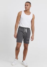 Shorts de algodón grises con cintura elástica y cordón blanco, con un logo impreso. Combinados con una camiseta sin mangas blanca y zapatillas blancas.