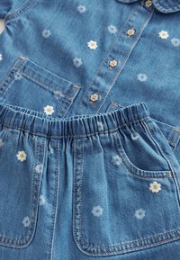 Tenue en denim bleu avec imprimés marguerites blanches, taille élastique sur le pantalon, chemise boutonnée avec boutons en forme de fleurs et poches avant.