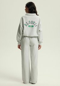 Szara, krótka bluza z napisem "BLOOVII 1999 CAMPUS DREAMER" w różowym i zielonym kolorze, w zestawie z pasującymi szarymi spodniami o szerokich nogawkach. Miękka tekstura materiału.