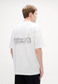 Bílý bavlněný tričko s lesklým, vyvýšeným logem 'HUGO' na zadní straně. Krátké rukávy, rovný lem a volný střih.