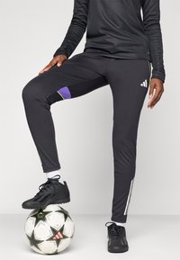 Svart treningbukser med lilla og grønne detaljer, med en slim fit, tre hvite striper på siden og en teksturert overflate.