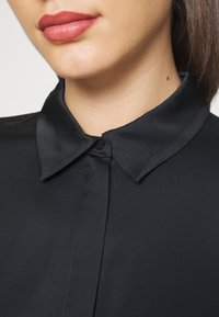 Femme portant une chemise noire boutonnée à col, montrant ses lèvres, son menton et son cou avec un petit grain de beauté sur le côté droit.