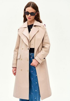 Kobieta w beżowym płaszczu trencz, czarnych okularach przeciwsłonecznych, czarnej bluzce i niebieskich dżinsach z małym kwiatowym haftem na jednolitym tle.