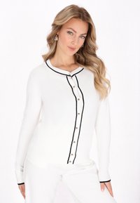 Weißer Strickcardigan mit schwarzem Besatz, runder Ausschnitt und goldenen Knopfdetails an der Vorderseite. Lange Ärmel mit schwarzen Bündchen.