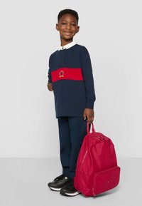 Navy langermet polo med rødstripet detalj, hvit krage, mørkeblå bukser og en rosa ryggsekk med glidelåser og frontlomme.