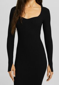 Femme portant une robe noire moulante à manches longues côtelées avec un décolleté carré, debout devant un fond clair uni.