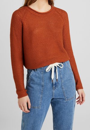 Rdzawego koloru dziergany, długi sweter z krótkim krojem, zestawiony z wysokim stanem niebieskich dżinsów z białym paskiem ze sznurkiem.